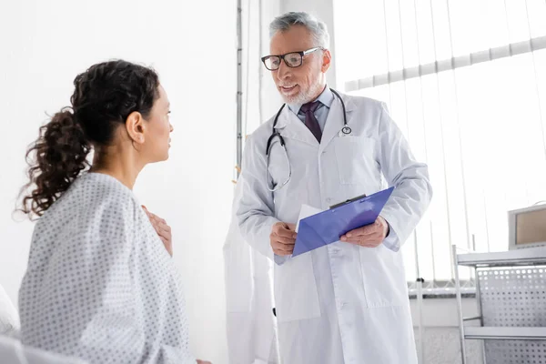 Sonriente médico sosteniendo portapapeles mientras habla con la mujer afroamericana en la clínica - foto de stock