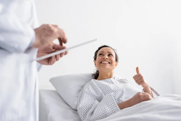 Mujer afroamericana feliz mostrando el pulgar hacia arriba cerca del médico con la tableta digital en primer plano borrosa - foto de stock