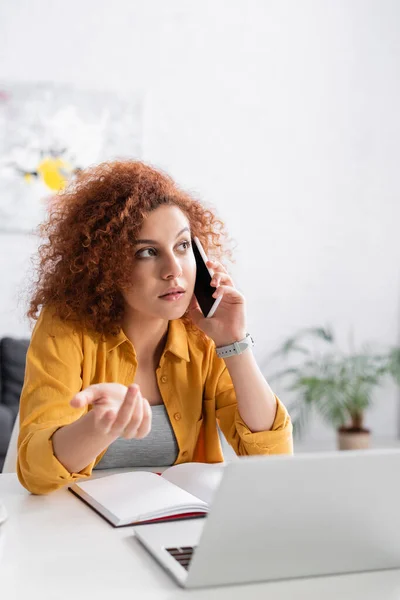 Thoughtful freelancer gesturing while talking on cellphone near laptop on blurred foreground — Stock Photo