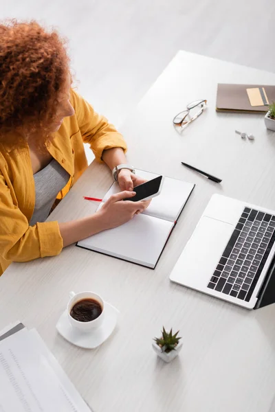 Vista de ángulo alto de la mensajería freelancer rizado en el teléfono inteligente cerca de la computadora portátil y la taza de café - foto de stock