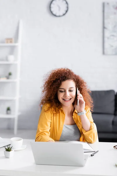 Sonriente freelancer llamando en smartphone cerca del ordenador portátil en casa - foto de stock