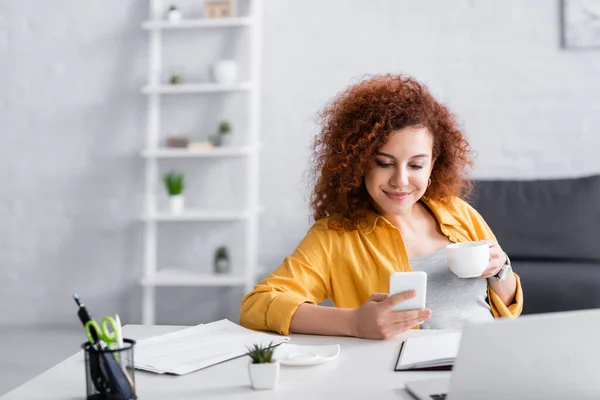 Glückliche Freelancer-Nachrichten auf dem Handy, während sie mit Kaffeetasse am Schreibtisch sitzen — Stockfoto