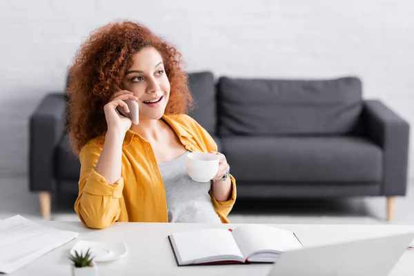 Joyful freelancer holding cup of coffee while talking on mobile phone at home — Stock Photo