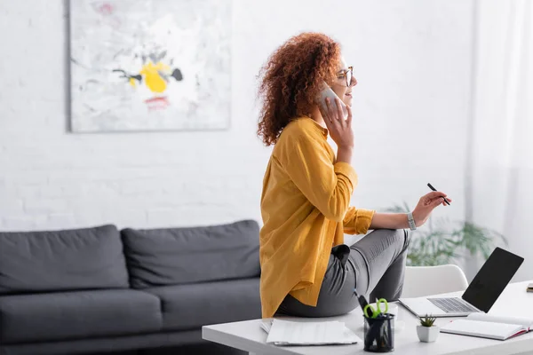 Joven freelancer hablando en smartphone mientras está sentado en el escritorio cerca de la computadora portátil con pantalla en blanco - foto de stock