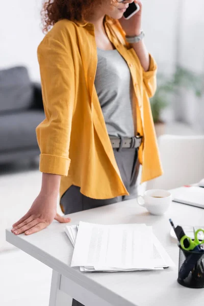 Partial view of freelancer talking on cellphone while standing near documents on desk — Stock Photo