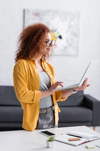 Curly freelancer in eyeglasses typing on laptop while standing near desk at home — Stock Photo