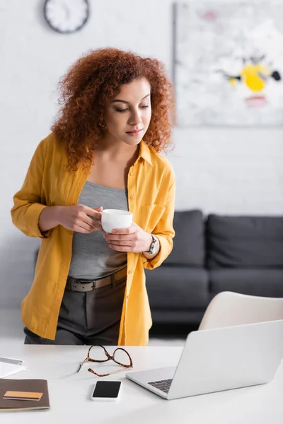 Junge Freiberuflerin hält Kaffeetasse in der Hand, während sie auf den Laptop am Schreibtisch blickt — Stockfoto