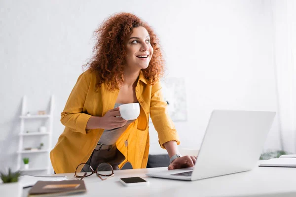 Alegre freelancer de pie con taza de café cerca de la computadora portátil en primer plano borrosa - foto de stock