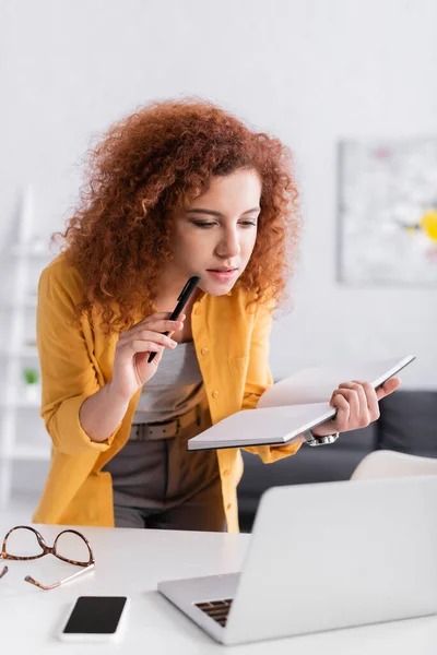 Thoughtful freelancer holding notebook and pen near laptop and mobile phone on blurred foreground — Stock Photo