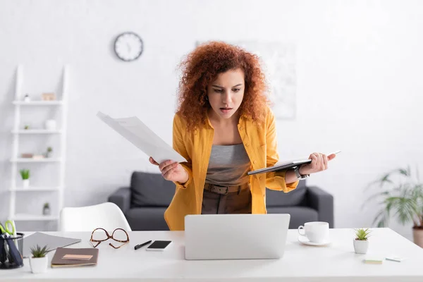 Serious freelancer holding documents and notebook while looking at laptop — Stock Photo