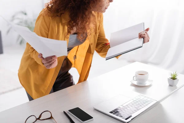 Cropped view of freelancer holding documents and notebook near laptop and mobile phone on desk — Stock Photo