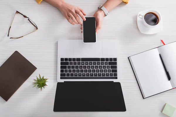 Partial view of teleworker holding smartphone with blank screen near laptop and coffee cup — Stock Photo