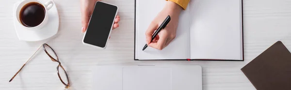 Vista recortada de freelancer segurando caneta e smartphone perto de notebook vazio, banner — Fotografia de Stock