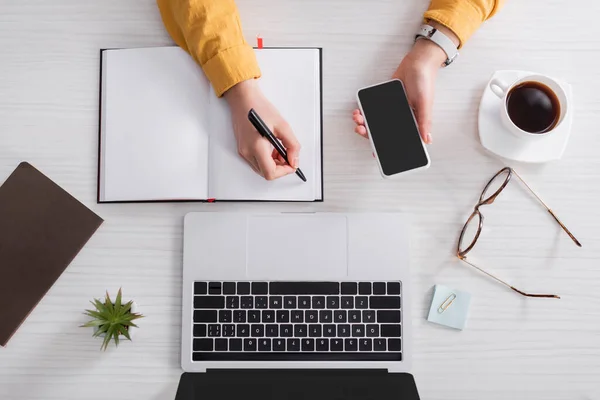 Vista recortada del freelancer que sostiene el teléfono inteligente con la pantalla en blanco y la escritura en el portátil cerca del ordenador portátil - foto de stock