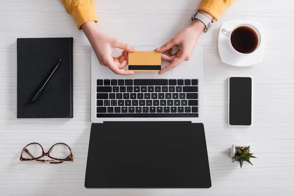 Vista parcial de la mujer que sostiene la tarjeta de crédito cerca del ordenador portátil y el teléfono celular con pantalla en blanco - foto de stock