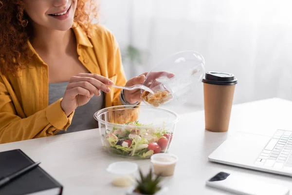 Partial view of smiling teleworker holding plastic fork near fresh salad and takeaway drink — Stock Photo