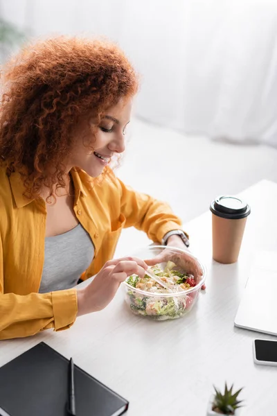 Telelavoro sorridente mangiare insalata da asporto vicino tazza di carta con caffè a casa — Foto stock