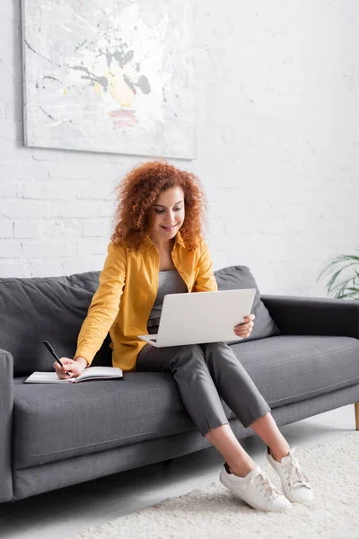 Happy freelancer holding pen near notebook while sitting on sofa with laptop — Stock Photo