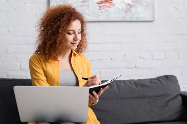 Hübsche Telearbeiterin schreibt in Notizbuch, während sie mit Laptop auf der Couch sitzt — Stockfoto