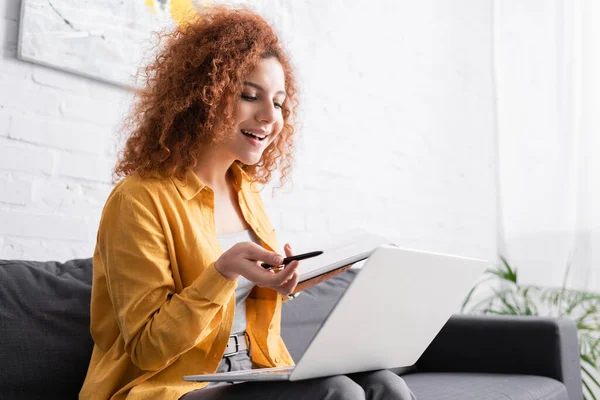 Joyful freelancer pointing with pen at laptop while holding notebook on sofa — Stock Photo