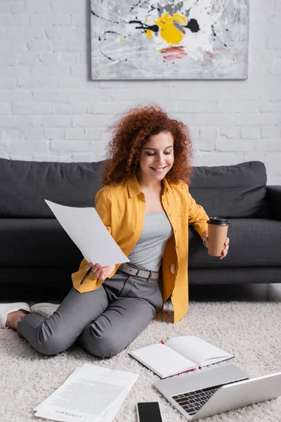 Smiling freelancer holding coffee to go and document while sitting on floor near laptop — Stock Photo