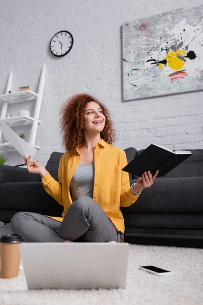 Glücklicher Freiberufler, der Notizbuch und Dokument in der Hand hält, während er auf dem Boden in Laptopnähe im unscharfen Vordergrund arbeitet — Stockfoto