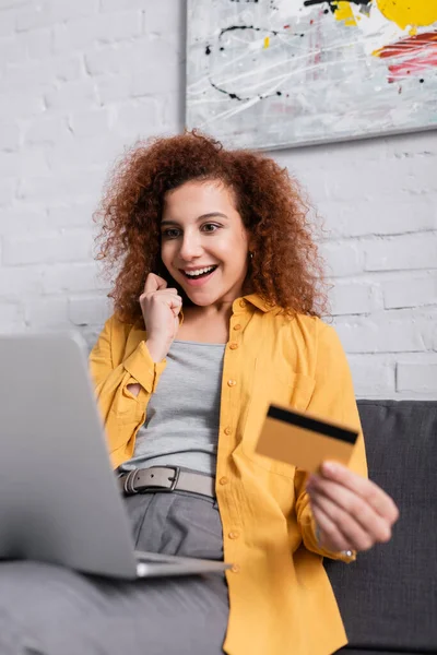 Femme heureuse tenant carte de crédit près d'un ordinateur portable sur le premier plan flou — Photo de stock