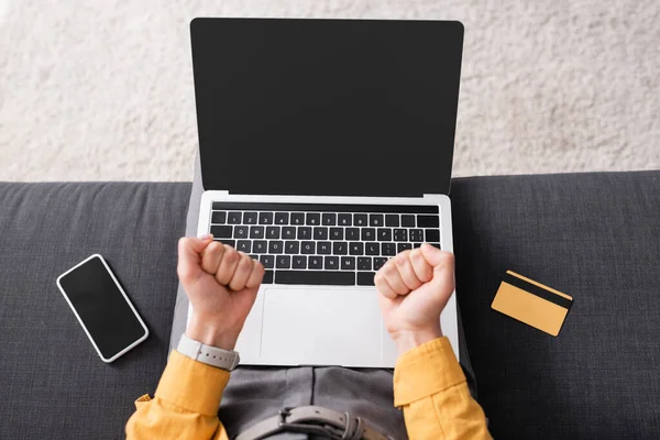 Partial view of woman with clenched fists sitting on sofa with laptop, smartphone and credit card — Stock Photo