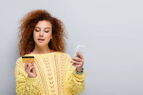 Кудрявая женщина в трикотажном свитере, держащая смартфон и кредитную карту изолированными на сером — стоковое фото