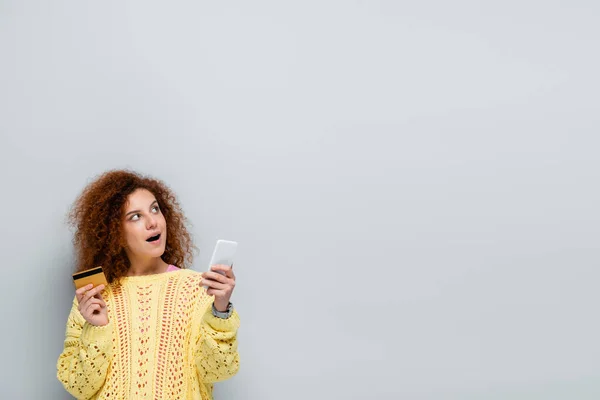 Donna sorpresa guardando lontano mentre tiene smartphone e carta di credito isolati su grigio — Foto stock