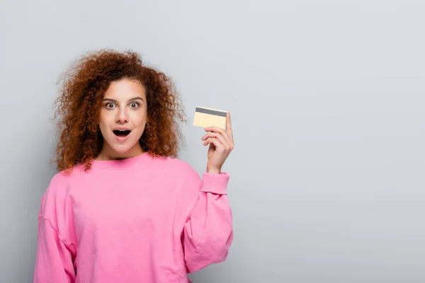 Donna sorpresa guardando la fotocamera mentre tiene la carta di credito isolata su grigio — Foto stock