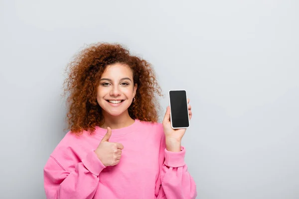 Femme heureuse montrant pouce vers le haut tout en tenant le téléphone portable avec écran vide isolé sur gris — Photo de stock