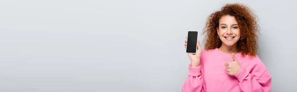 Mujer alegre mostrando el pulgar hacia arriba mientras sostiene el teléfono celular con pantalla en blanco aislado en gris, pancarta - foto de stock