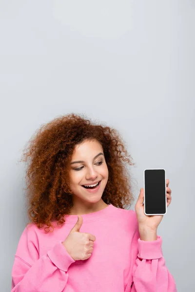 Gioiosa donna in possesso di smartphone con schermo bianco e mostrando pollice in alto isolato sul grigio — Foto stock