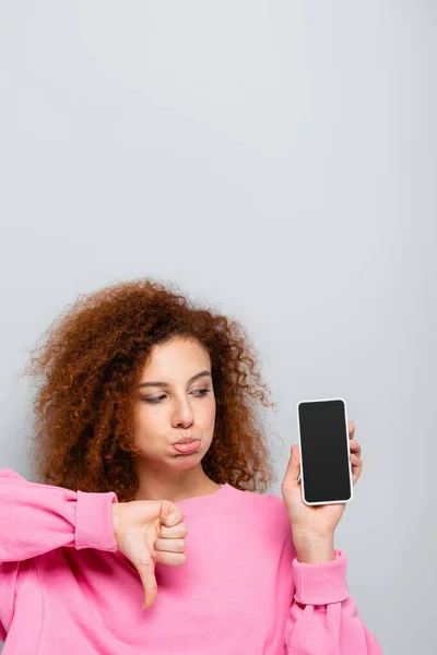 Mujer disgustada haciendo pucheros labios y mostrando el pulgar hacia abajo cerca de teléfono inteligente con pantalla en blanco aislado en gris - foto de stock