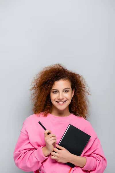 Curly woman with notebook and pen smiling at camera isolated on grey — Stock Photo