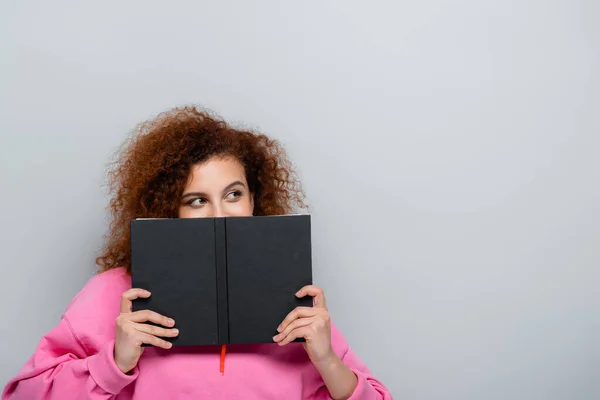 Young woman with curly hair obscuring face with notebook isolated on grey — Stock Photo