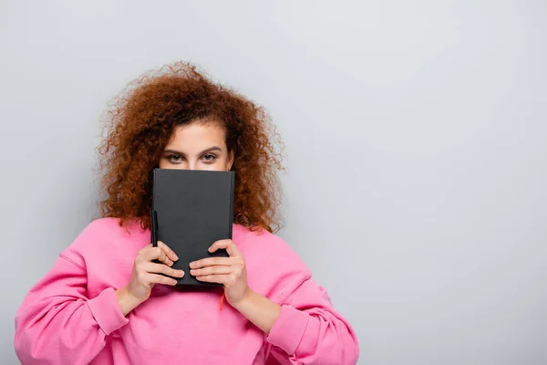 Mulher encaracolado obscurecendo rosto com notebook enquanto olha para a câmera isolada em cinza — Fotografia de Stock