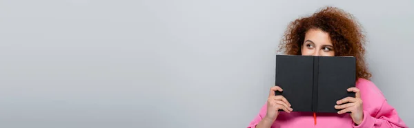 Young woman with wavy hair obscuring face with notebook isolated on grey, banner — Stock Photo