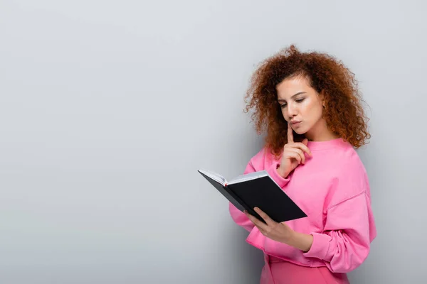 Mujer rizada sosteniendo el cuaderno y tocando la cara mientras piensa en fondo gris - foto de stock