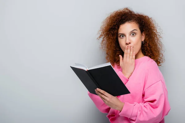 Schockierte Frau bedeckt Mund mit der Hand, während sie Notizbuch auf grauem Hintergrund hält — Stockfoto