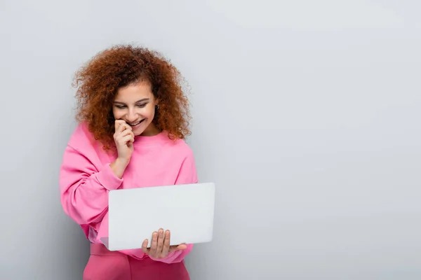 Femme gaie tenant la main près du visage tout en regardant ordinateur portable sur fond gris — Photo de stock