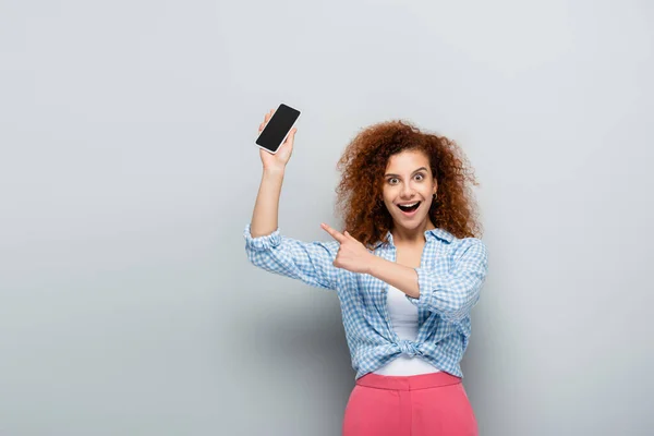 Mujer asombrada señalando con el dedo mientras sostiene el teléfono inteligente con pantalla en blanco sobre fondo gris — Stock Photo