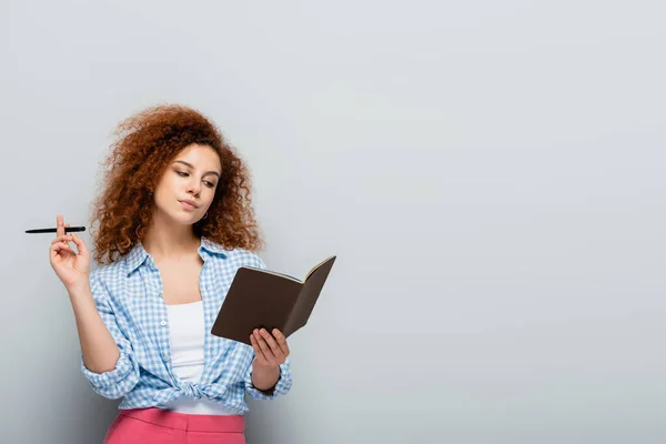 Thoughtful woman in plaid shirt holding pen and notebook on grey background — Stock Photo