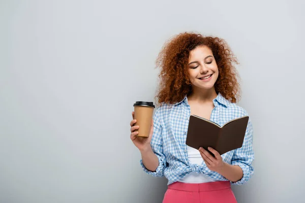 Cheerful woman looking in notebook while holding paper cup on grey background — Stock Photo