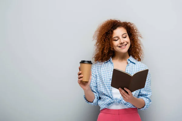 Mujer alegre mirando en cuaderno mientras sostiene la taza de papel sobre fondo gris - foto de stock