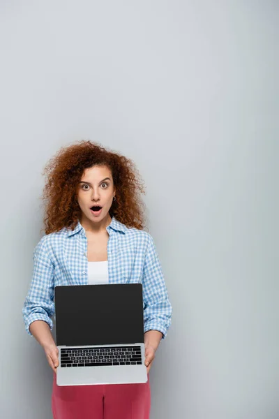 Shocked woman looking at camera while showing laptop with blank screen on grey background — Stock Photo