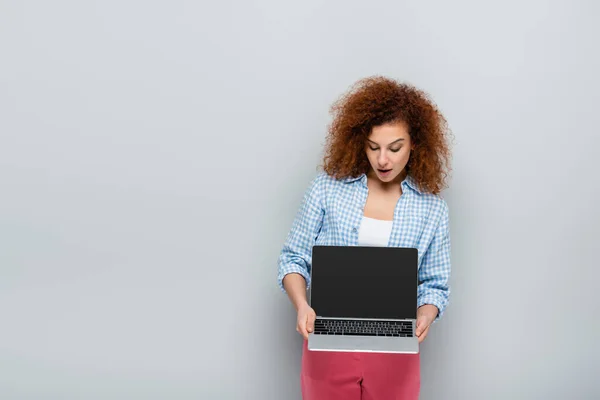 Donna sorpresa con i capelli ricci che tengono il computer portatile con schermo bianco su sfondo grigio — Foto stock