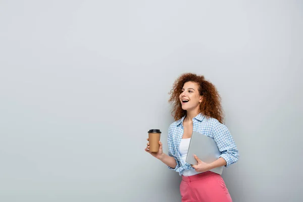 Femme incroyable portant du café pour aller et ordinateur portable sur fond gris — Photo de stock