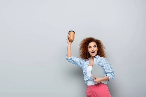 Excited woman holding coffee to go while carrying laptop on grey background — Stock Photo